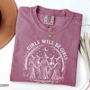 K&ouml;nnte beinhalten: Beerenfarbenes T-Shirt mit dem wei&szlig;en Text "GIRLS WILL BE GIRLS". Die Grafik zeigt f&uuml;nf Hexenfiguren, die sich unter einem Mond und Sternen an den H&auml;nden halten. Eine silberne Halskette mit Anh&auml;nger liegt auf dem Shirt.