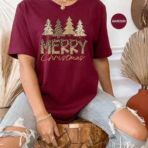 May include: A maroon t-shirt with a graphic of three Christmas trees decorated with leopard print and gold glitter. The text "Merry Christmas" is printed below the trees.