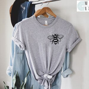 May include: A gray t-shirt with a black bee graphic on the front. The shirt is hanging on a hanger and is tied in a knot at the bottom.