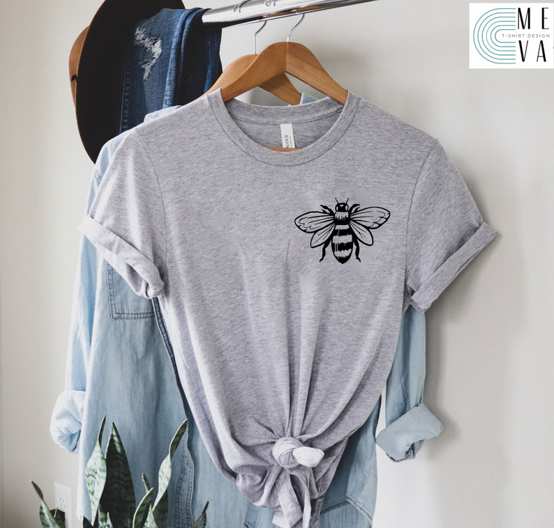 May include: A gray t-shirt with a black bee graphic on the front. The shirt is hanging on a hanger and is tied in a knot at the bottom.
