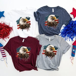 May include: Four t-shirts with a graphic of a bald eagle in front of an American flag with paint splatters. The shirts are in white, navy blue, maroon, and heather gray.