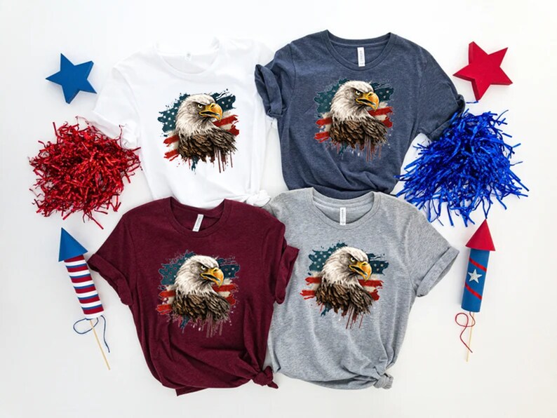 May include: Four t-shirts with a graphic of a bald eagle in front of an American flag with paint splatters. The shirts are in white, navy blue, maroon, and heather gray.