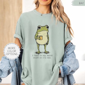 May include: A light green t-shirt featuring a cartoon frog holding a mug. The frog is green and beige, with large eyes and a coffee mug. The text "i have to say weird stuff or i'll die" is printed below the frog.