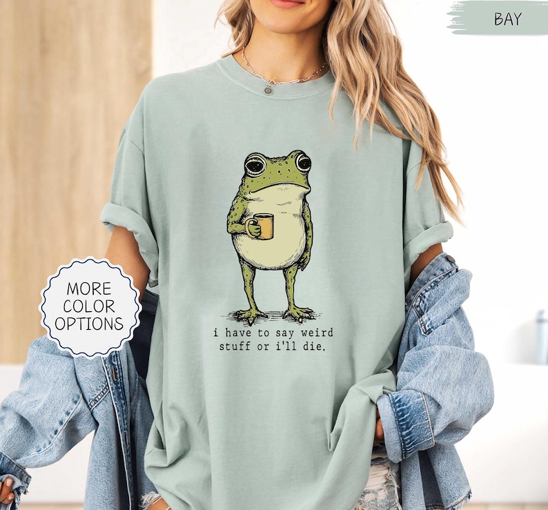May include: A light green t-shirt featuring a cartoon frog holding a mug. The frog is green and beige, with large eyes and a coffee mug. The text "i have to say weird stuff or i'll die" is printed below the frog.