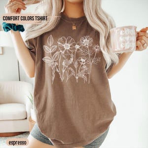May include: Espresso-colored Comfort Colors t-shirt featuring a white floral design. The shirt has short sleeves and a crew neck. The text "COMFORT COLORS TSHIRT" is printed on the top left of the shirt.