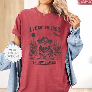 May include: Crimson-colored t-shirt with the text "I've Got Friends in Low Places" and a graphic of a corgi wearing a cowboy hat, bandana, and surrounded by cacti. The shirt also has the words "More Color Options" on a white label.