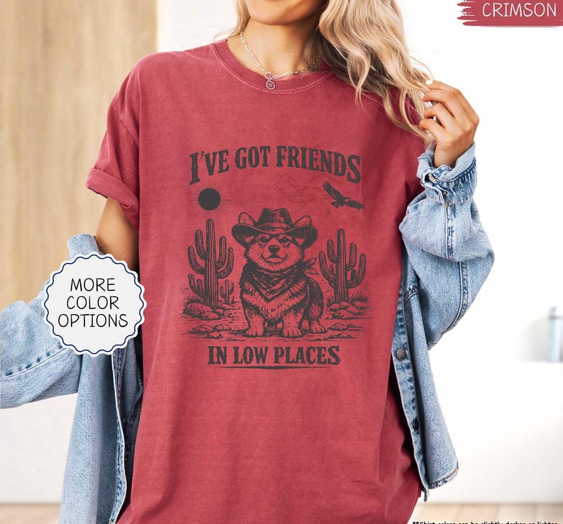 May include: Crimson-colored t-shirt with the text "I've Got Friends in Low Places" and a graphic of a corgi wearing a cowboy hat, bandana, and surrounded by cacti. The shirt also has the words "More Color Options" on a white label.