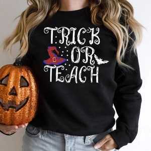 Puede incluir: Sudadera negra con las palabras "TRICK OR TEACH" en escritura blanca. Un sombrero de bruja en rojo, azul y negro aparece en el dise&ntilde;o, junto con estrellas y un murci&eacute;lago. Una calabaza naranja brillante est&aacute; en la imagen.