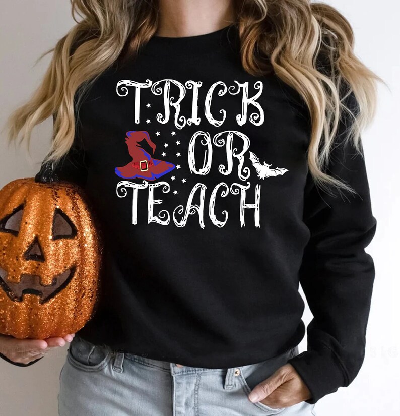 Puede incluir: Sudadera negra con las palabras "TRICK OR TEACH" en escritura blanca. Un sombrero de bruja en rojo, azul y negro aparece en el dise&ntilde;o, junto con estrellas y un murci&eacute;lago. Una calabaza naranja brillante est&aacute; en la imagen.