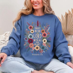 Peut inclure: Un sweat-shirt bleu avec un motif floral et le texte "Trust in the Lord with all your heart" et "Proverbes 3:5". Le motif floral comprend des fleurs dans les tons de rouge, rose, jaune et bleu.