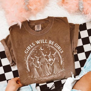 K&ouml;nnte beinhalten: Ein braunes T-Shirt mit dem Text "GIRLS WILL BE GIRLS" in einem wei&szlig;en, kreisf&ouml;rmigen Design. Das Design zeigt f&uuml;nf Hexenfiguren in Kleidern und H&uuml;ten, mit einem Mond und Sternen. Das Shirt hat kurze &Auml;rmel und einen Rundhalsausschnitt.