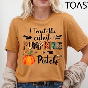 May include: Mustard-coloured t-shirt with the text "I Teach the Cutest Pumpkins in the Patch." The design includes a colourful pumpkin graphic and autumn leaves. The shirt is a comfortable, casual style.
