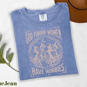 K&ouml;nnte beinhalten: Blaues Jeans-T-Shirt mit dem Text "GOD FORBID WOMEN HAVE HOBBIES" in einem kreisf&ouml;rmigen Design mit drei Hexen. Das Shirt ist von der Marke Comfort Colors und liegt gefaltet auf einer wei&szlig;en Oberfl&auml;che.