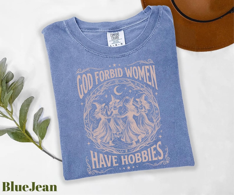 K&ouml;nnte beinhalten: Blaues Jeans-T-Shirt mit dem Text "GOD FORBID WOMEN HAVE HOBBIES" in einem kreisf&ouml;rmigen Design mit drei Hexen. Das Shirt ist von der Marke Comfort Colors und liegt gefaltet auf einer wei&szlig;en Oberfl&auml;che.