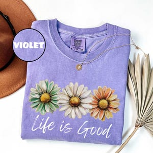 May include: Violet-colored sweatshirt with a crew neck and the words "Life is Good" in white script. The shirt features a floral design of three daisies in green, white, and orange. A gold necklace with a pendant is draped on the shirt.