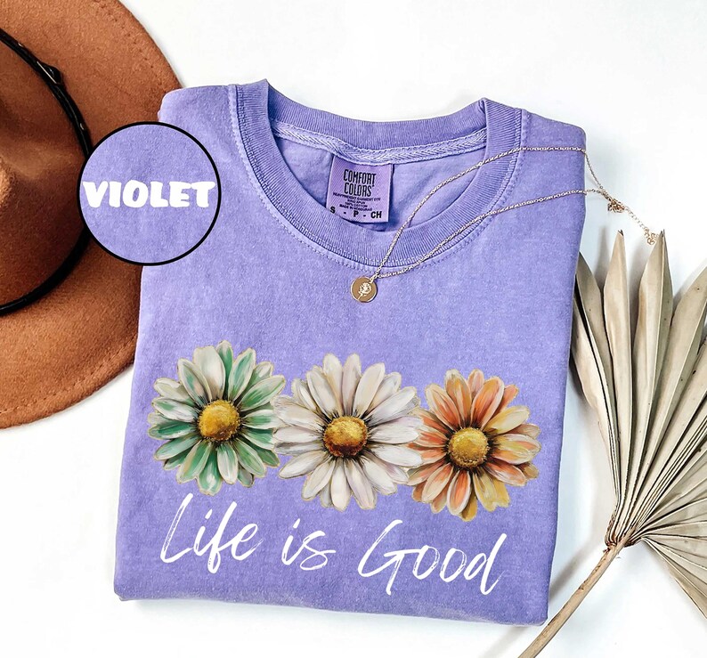 May include: Violet-colored sweatshirt with a crew neck and the words "Life is Good" in white script. The shirt features a floral design of three daisies in green, white, and orange. A gold necklace with a pendant is draped on the shirt.