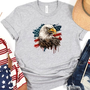 May include: A gray t-shirt with a graphic of a bald eagle in front of an American flag with paint splatters. The eagle's head is turned to the right and its beak is open.