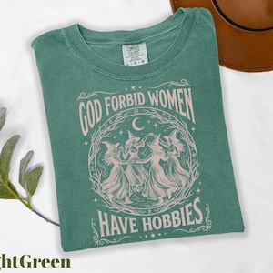 K&ouml;nnte beinhalten: Hellgr&uuml;nes T-Shirt mit dem Text "GOD FORBID WOMEN HAVE HOBBIES" in einem kreisf&ouml;rmigen Design mit tanzenden Hexen. Das Shirt ist von der Marke Comfort Colors und das Design ist hellrosa.
