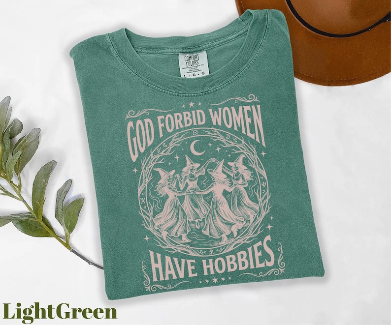 K&ouml;nnte beinhalten: Hellgr&uuml;nes T-Shirt mit dem Text "GOD FORBID WOMEN HAVE HOBBIES" in einem kreisf&ouml;rmigen Design mit tanzenden Hexen. Das Shirt ist von der Marke Comfort Colors und das Design ist hellrosa.