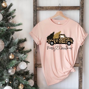 Op de afbeelding: Een roze T-shirt met een zwarte en gouden vrachtwagen die een kerstboom vervoert. De vrachtwagen is versierd met een luipaardprint. De tekst "Merry Christmas" staat onder de vrachtwagen.