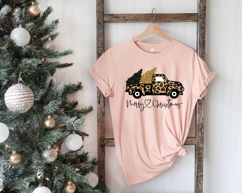 Op de afbeelding: Een roze T-shirt met een zwarte en gouden vrachtwagen die een kerstboom vervoert. De vrachtwagen is versierd met een luipaardprint. De tekst "Merry Christmas" staat onder de vrachtwagen.
