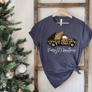 Op de afbeelding: Een donkerblauw T-shirt met een afbeelding van een vrachtwagen die een kerstboom vervoert. De vrachtwagen is versierd met een luipaardprint en gouden glitter. De tekst "Merry Christmas" staat onder de vrachtwagen.