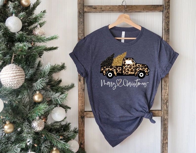 Op de afbeelding: Een donkerblauw T-shirt met een afbeelding van een vrachtwagen die een kerstboom vervoert. De vrachtwagen is versierd met een luipaardprint en gouden glitter. De tekst "Merry Christmas" staat onder de vrachtwagen.