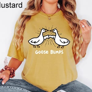 May include: A mustard yellow t-shirt featuring a graphic of two geese facing each other with the text "Goose Bumps" underneath.