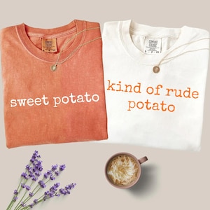 May include: Two t-shirts, one orange and one white, with the phrases "sweet potato" and "kind of rude potato" printed on them. Each shirt has a gold necklace. A cup of coffee and lavender sprigs are also in the image.