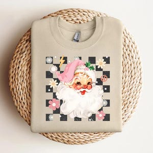May include: Beige sweatshirt featuring a retro Santa Claus graphic. The design includes a pink Santa hat, a white beard, and a checkered background with flowers and a lightning bolt. A peace sign is also included in the design.
