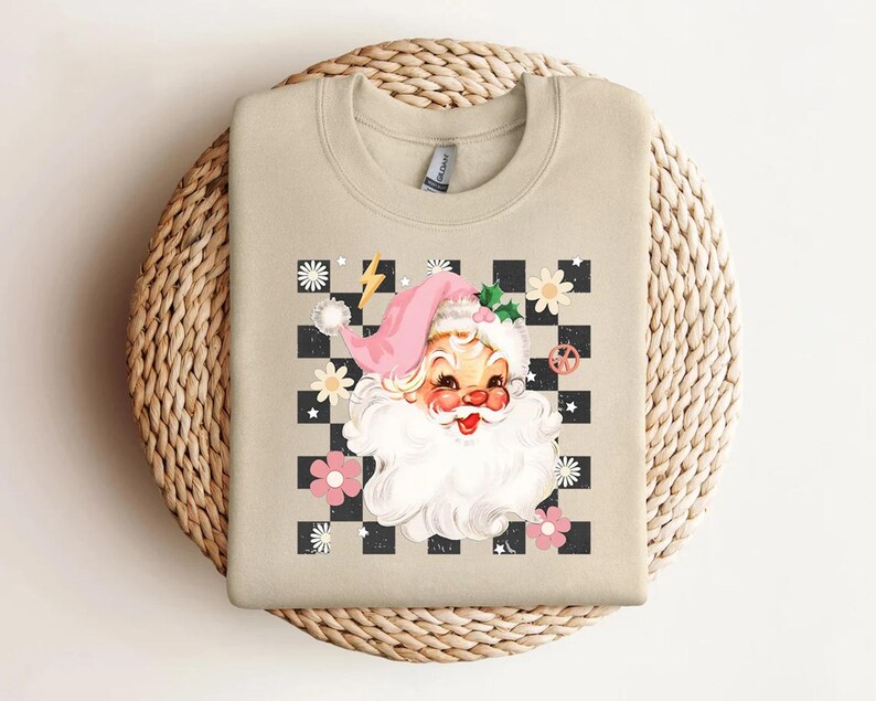 May include: Beige sweatshirt featuring a retro Santa Claus graphic. The design includes a pink Santa hat, a white beard, and a checkered background with flowers and a lightning bolt. A peace sign is also included in the design.
