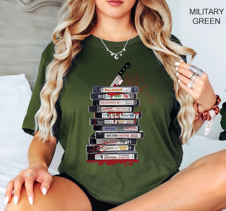 Pode incluir: T-shirt verde militar com um desenho de uma pilha de fitas VHS com t&iacute;tulos de filmes de terror e uma faca ensanguentada. Os t&iacute;tulos incluem "Halloween" e "Hellraiser". A camisa tem gola redonda e mangas curtas.
