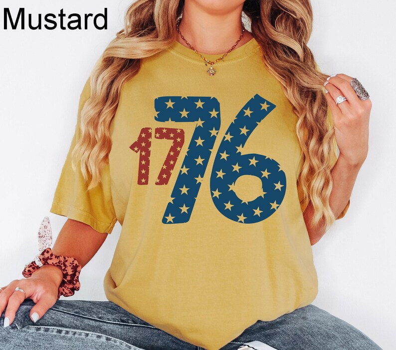 Puede incluir: Una camiseta amarilla mostaza con un gr&aacute;fico rojo y azul del n&uacute;mero 176 con estrellas. El gr&aacute;fico est&aacute; formado por estrellas rojas y azules sobre un fondo blanco.