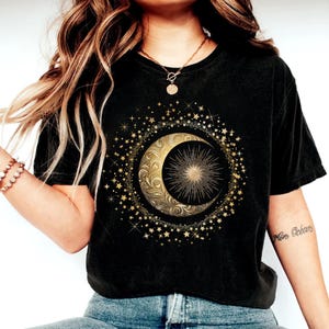 May include: Black t-shirt featuring a gold crescent moon and sunburst design, surrounded by stars. The celestial graphic is centered on the shirt. The shirt is made of a soft material.