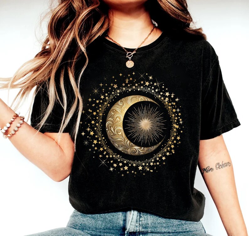 May include: Black t-shirt featuring a gold crescent moon and sunburst design, surrounded by stars. The celestial graphic is centered on the shirt. The shirt is made of a soft material.