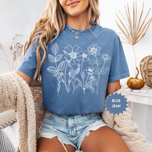 May include: Blue jean colored t-shirt with a white floral design. The t-shirt has a crew neck and short sleeves. The text "BLUE JEAN" is on a blue starburst-shaped tag.