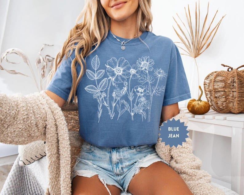 May include: Blue jean colored t-shirt with a white floral design. The t-shirt has a crew neck and short sleeves. The text "BLUE JEAN" is on a blue starburst-shaped tag.