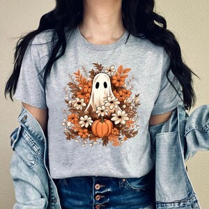 May include: A light gray t-shirt featuring a Halloween-themed graphic. The design includes a white ghost with black eyes surrounded by orange and white flowers, brown leaves, and a small orange pumpkin. The shirt is a casual wear item.