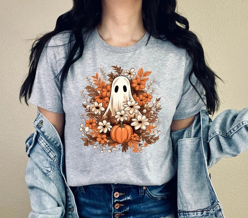 May include: A light gray t-shirt featuring a Halloween-themed graphic. The design includes a white ghost with black eyes surrounded by orange and white flowers, brown leaves, and a small orange pumpkin. The shirt is a casual wear item.