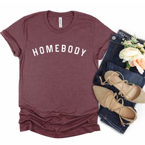 Puede incluir: Una camiseta de color granate con la palabra "HOMEBODY" impresa en blanco en la parte delantera. La camiseta se combina con unos vaqueros azules y unas sandalias beige.