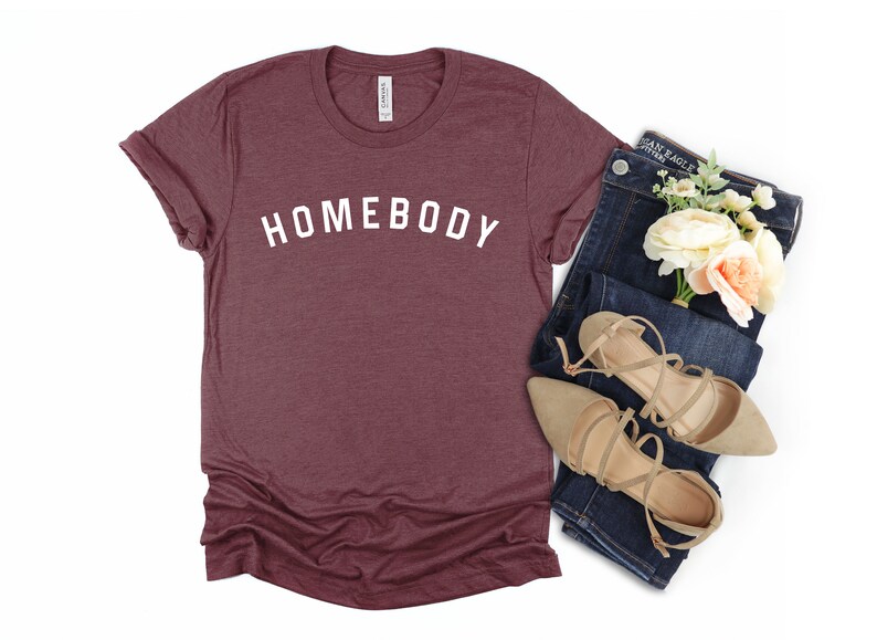 Puede incluir: Una camiseta de color granate con la palabra "HOMEBODY" impresa en blanco en la parte delantera. La camiseta se combina con unos vaqueros azules y unas sandalias beige.