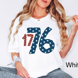 Puede incluir: Camiseta blanca con un gr&aacute;fico rojo, blanco y azul del n&uacute;mero 1776. Los n&uacute;meros est&aacute;n llenos de estrellas blancas sobre un fondo azul.