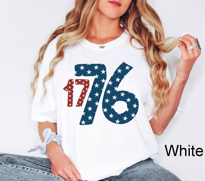 Puede incluir: Camiseta blanca con un gr&aacute;fico rojo, blanco y azul del n&uacute;mero 1776. Los n&uacute;meros est&aacute;n llenos de estrellas blancas sobre un fondo azul.