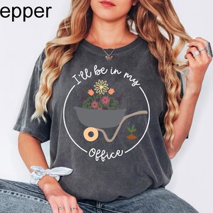 Op de afbeelding: Een donkergrijs T-shirt met een witte cirkelvormige afbeelding. De afbeelding toont een kruiwagen gevuld met kleurrijke bloemen en de tekst "I'll be in my office".