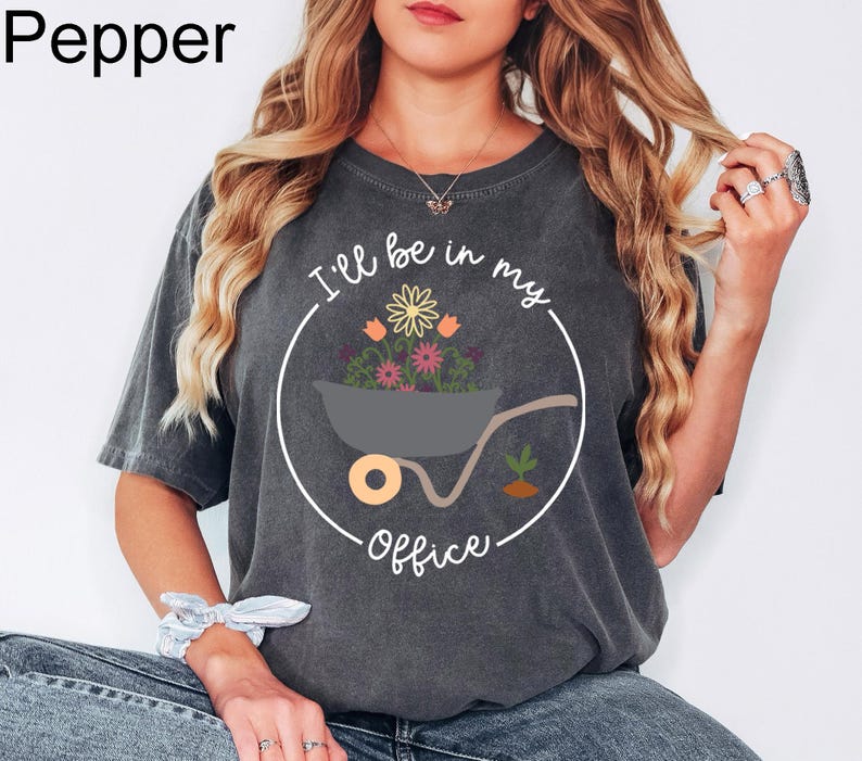 Op de afbeelding: Een donkergrijs T-shirt met een witte cirkelvormige afbeelding. De afbeelding toont een kruiwagen gevuld met kleurrijke bloemen en de tekst "I'll be in my office".