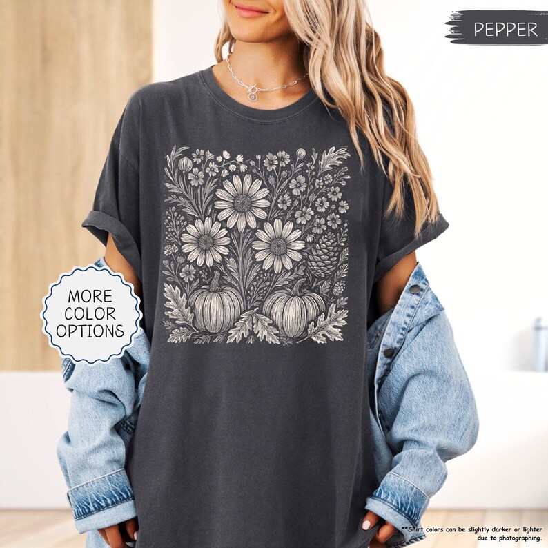 May include: Dark gray t-shirt featuring a floral and pumpkin design. The graphic includes large daisies, smaller flowers, and pumpkins. The shirt has rolled-up sleeves and is part of a fall-themed collection. The text "MORE COLOR OPTIONS" is visible.
