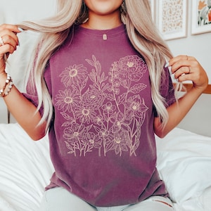 K&ouml;nnte beinhalten: Ein kurz&auml;rmeliges, pflaumenfarbenes T-Shirt mit einer detaillierten floralen Strichzeichnung in Hellbeige. Das Design umfasst verschiedene Blumen und Bl&auml;tter und schafft ein botanisches Thema. Das Shirt ist aus weichem Material.