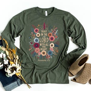 Peut inclure: Un t-shirt vert olive &agrave; manches longues avec un motif floral et le texte "Trust in the Lord with all your heart" et "Proverbes 3:5". Le t-shirt est associ&eacute; &agrave; un jean bleu, des chaussures beiges et un bouquet de fleurs.
