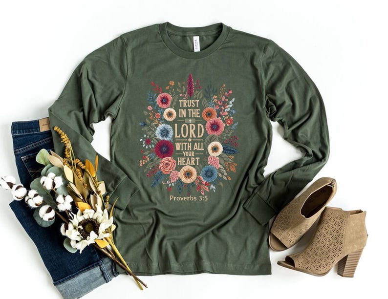 Peut inclure: Un t-shirt vert olive &agrave; manches longues avec un motif floral et le texte "Trust in the Lord with all your heart" et "Proverbes 3:5". Le t-shirt est associ&eacute; &agrave; un jean bleu, des chaussures beiges et un bouquet de fleurs.