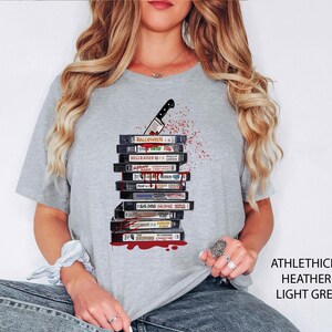 Pode incluir: T-shirt cinzenta clara com um design de cassetes VHS, uma faca e salpicos de sangue. As cassetes t&ecirc;m t&iacute;tulos de filmes como "Halloween" e "Hellraiser". O texto "ATHLETHICH HEATHER LIGHT GREY" est&aacute; impresso no lado direito.